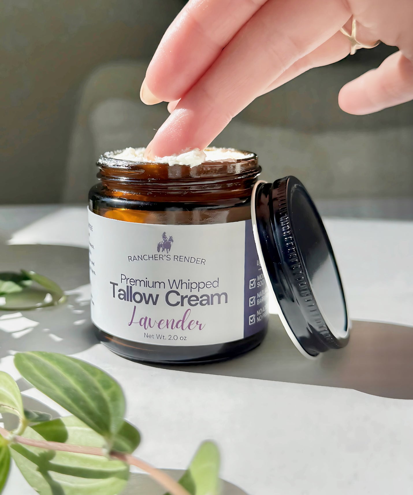 Hand applying lavender tallow cream from a jar labeled 'Rancher's Render Lavender Whipped Tallow' on a light surface with greenery.